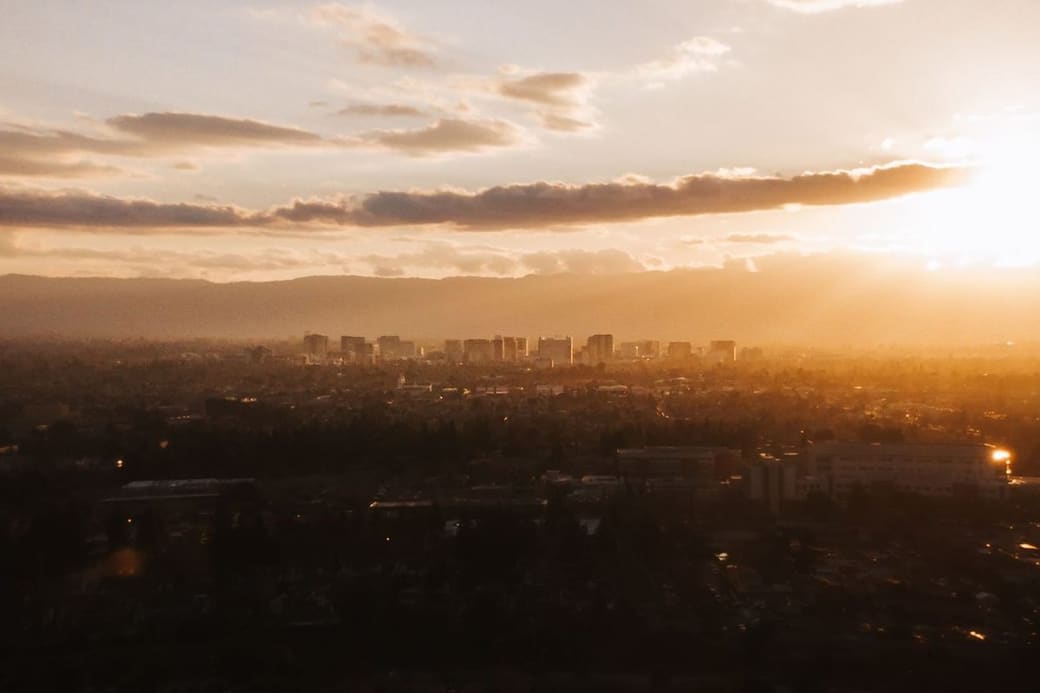 A low city skyline across a dark valley at sunset, the sun breaking through a band of cloud above distant hills