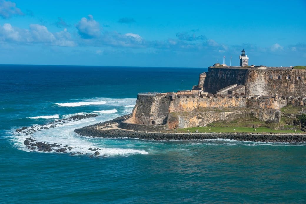 A massive stone coastal fortress with a small lighthouse sits on a headland above a curving rock breakwater, with white surf rolling in from a deep blue sea.