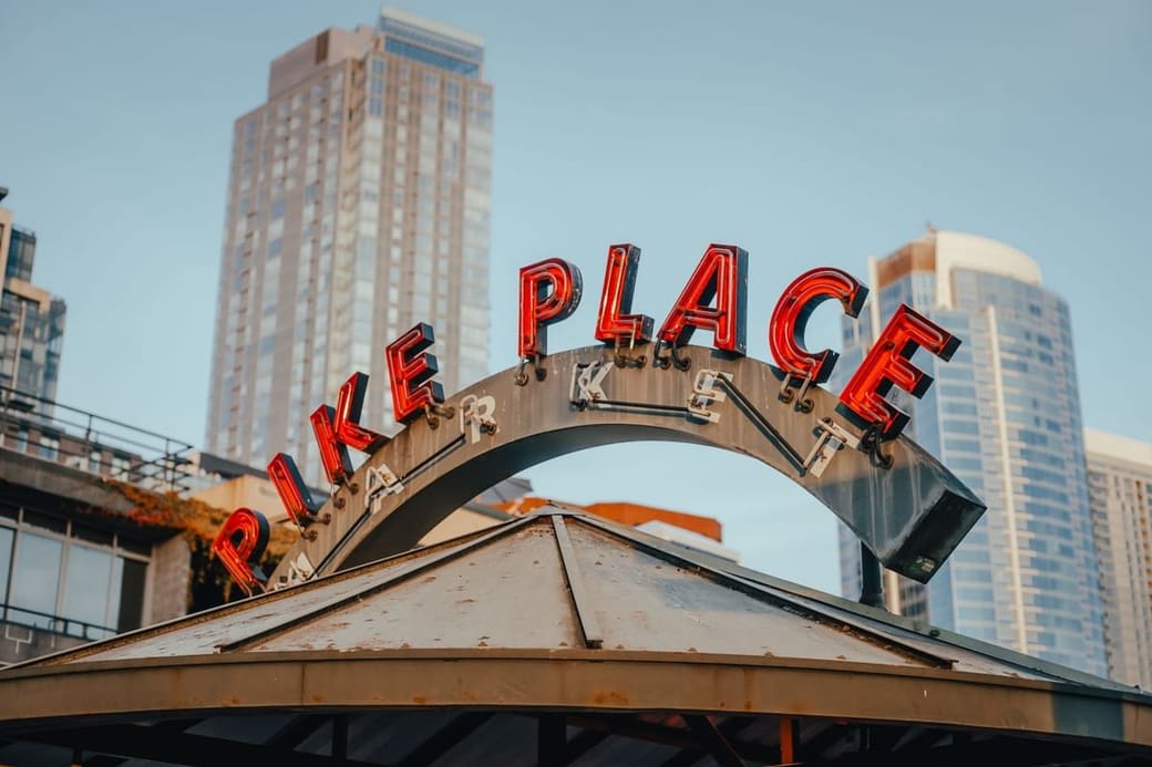 A curved metal arch over a street carrying a large illuminated sign reading Pike Place, with tall buildings rising behind it