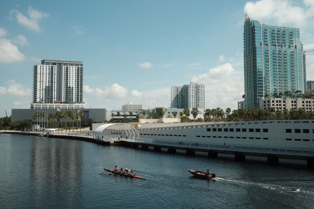 Rowing crews on flat water in daylight, with a white curved-roof building and glass towers along the far bank