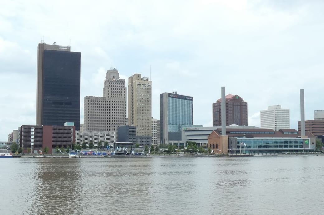 A downtown skyline seen flat across a wide river in daylight, a dark glass tower at the left and lower brick and stone blocks along the waterfront