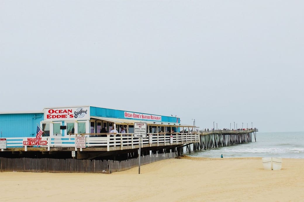 A blue and white seafood restaurant on a wooden pier stretching over a wide sandy beach into the sea, under a pale sky