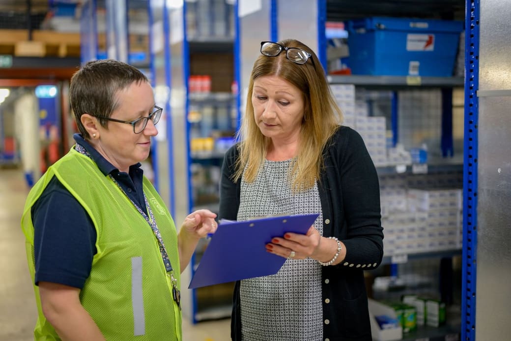 Two warehouse staff, one in a high-visibility vest, going through an inventory checklist on a clipboard beside stocked shelving