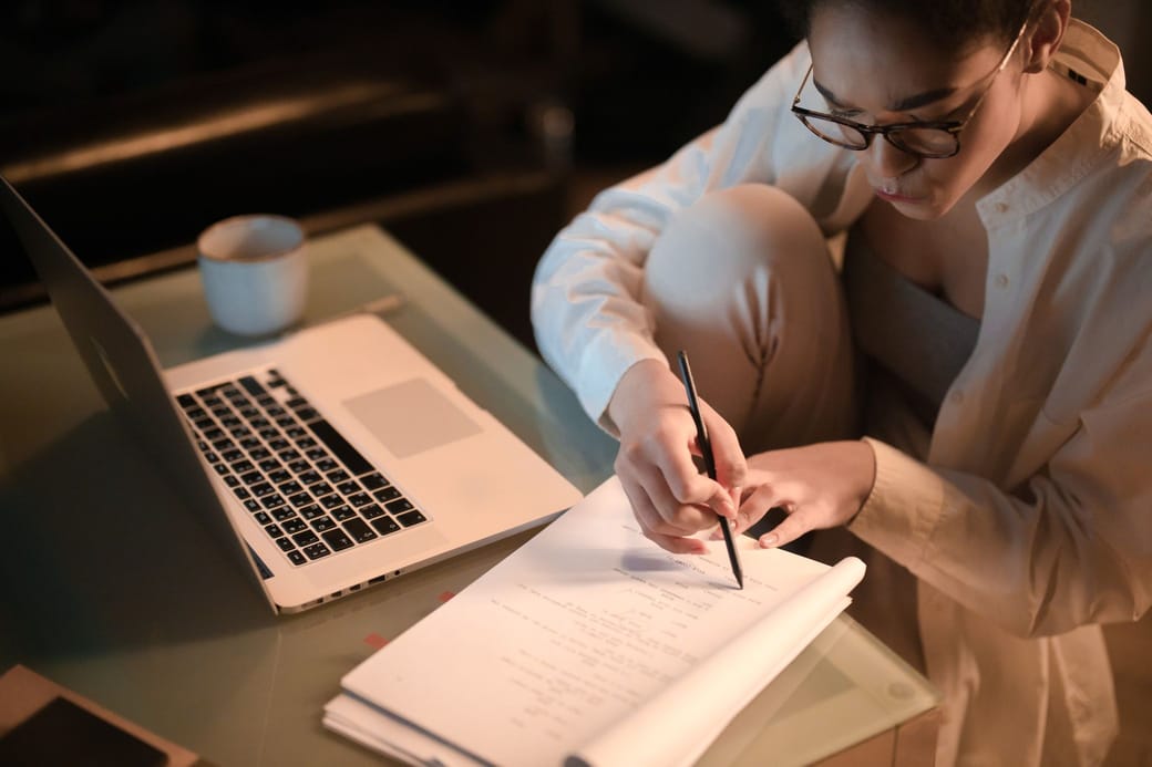A woman in glasses and a white shirt writing notes on printed pages beside an open laptop and a mug, lit by a warm lamp at night