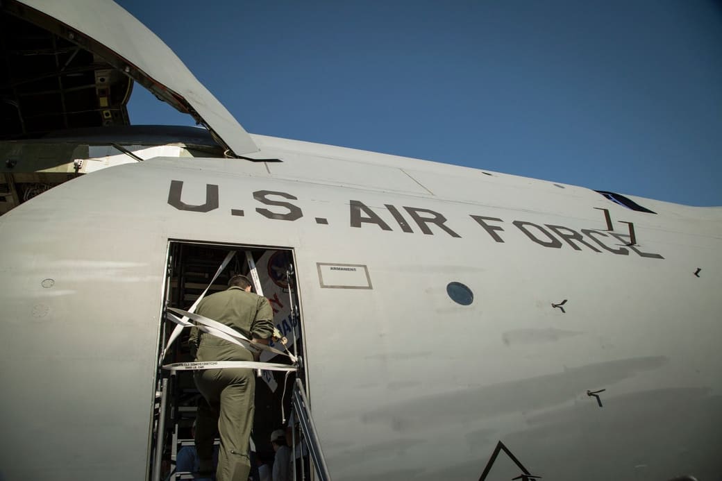 An airman in a flight suit climbing into the side door of a gray U.S. Air Force transport plane under a clear blue sky