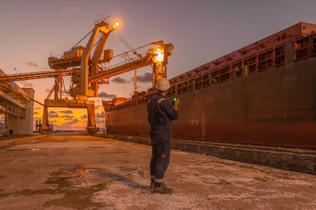 A dock worker in a hard hat watching a container ship being worked at a port terminal at dusk