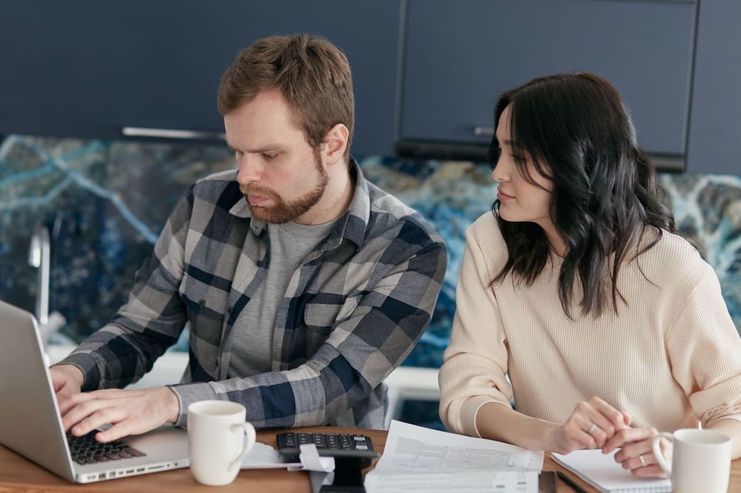 Two people at a kitchen table going over bills with a laptop, a calculator and printed statements
