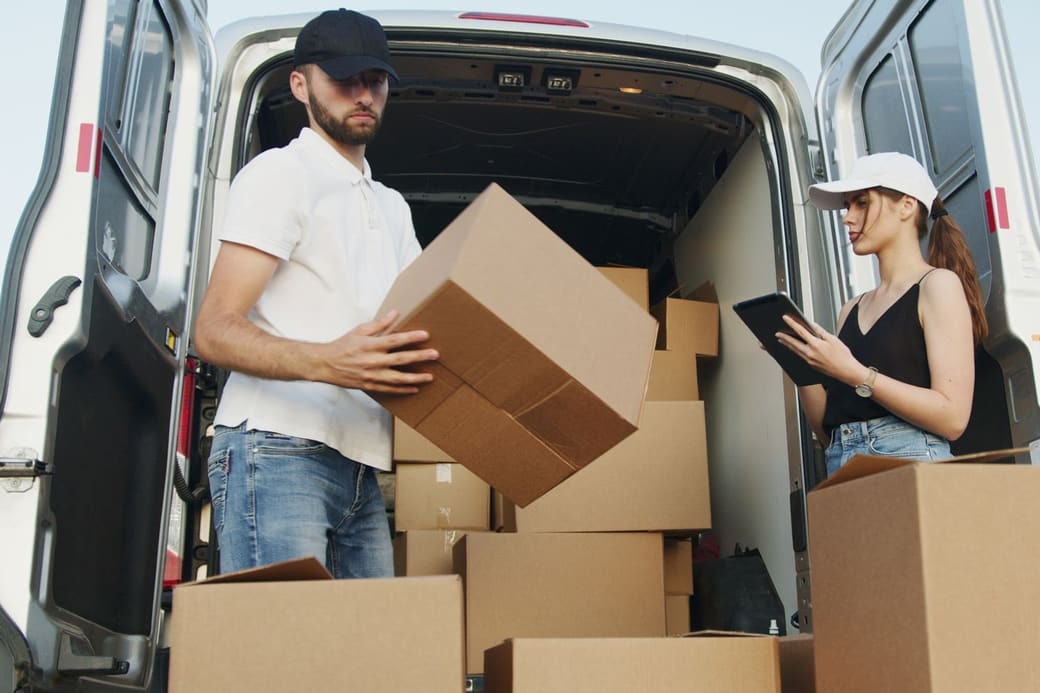 Two delivery workers loading cartons into a van, one carrying a box and one checking an order on a tablet