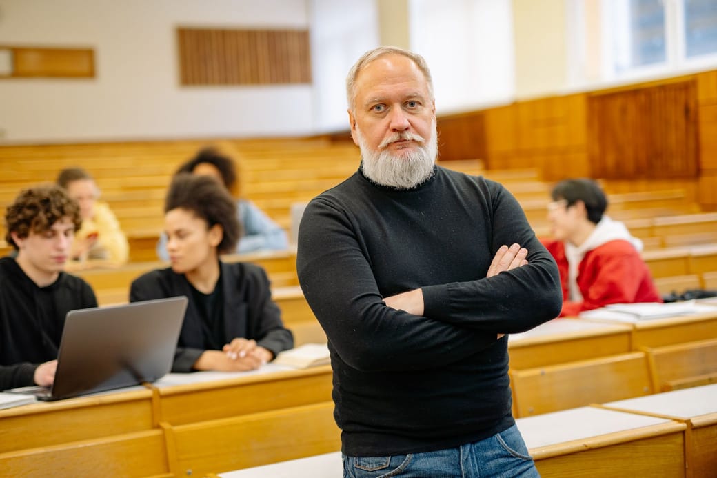 A gray-bearded professor in a black turtleneck standing with arms folded in a wood-paneled lecture hall, students working at the benches behind him