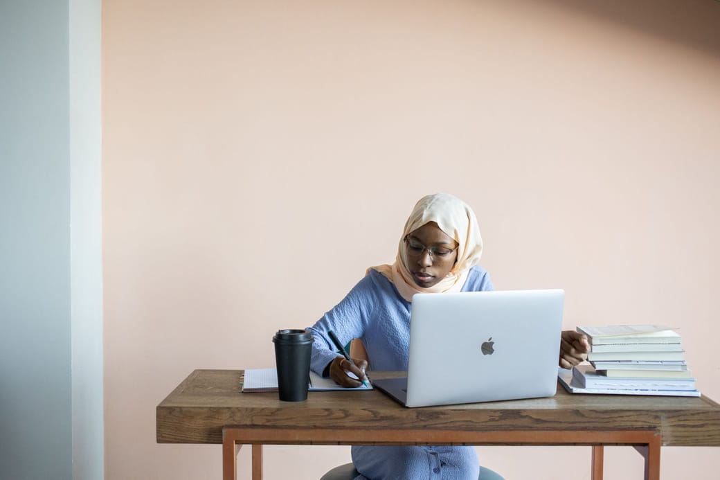 A woman in a peach headscarf and glasses taking notes beside an open laptop, a coffee cup and a stack of books at a wooden desk