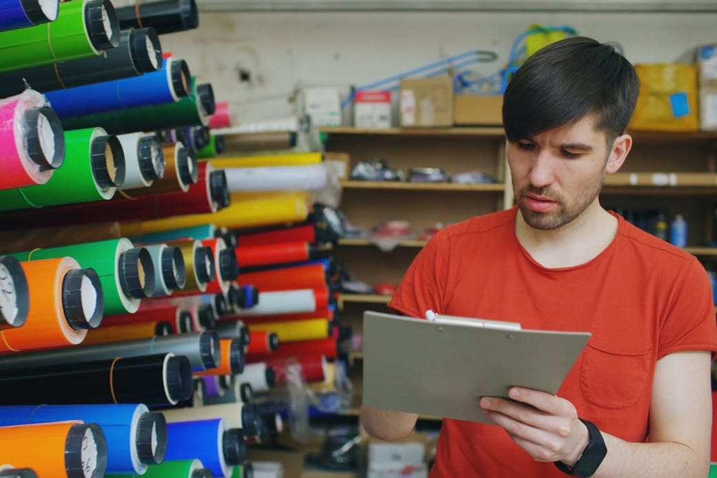A man in a red T-shirt checking a clipboard beside racks of colored material rolls in a stockroom