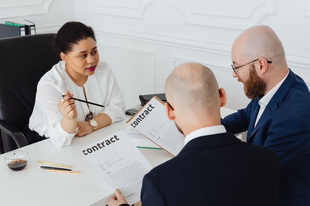 Three people at a white desk going over printed contracts, one of them pointing with a pencil as she explains a term