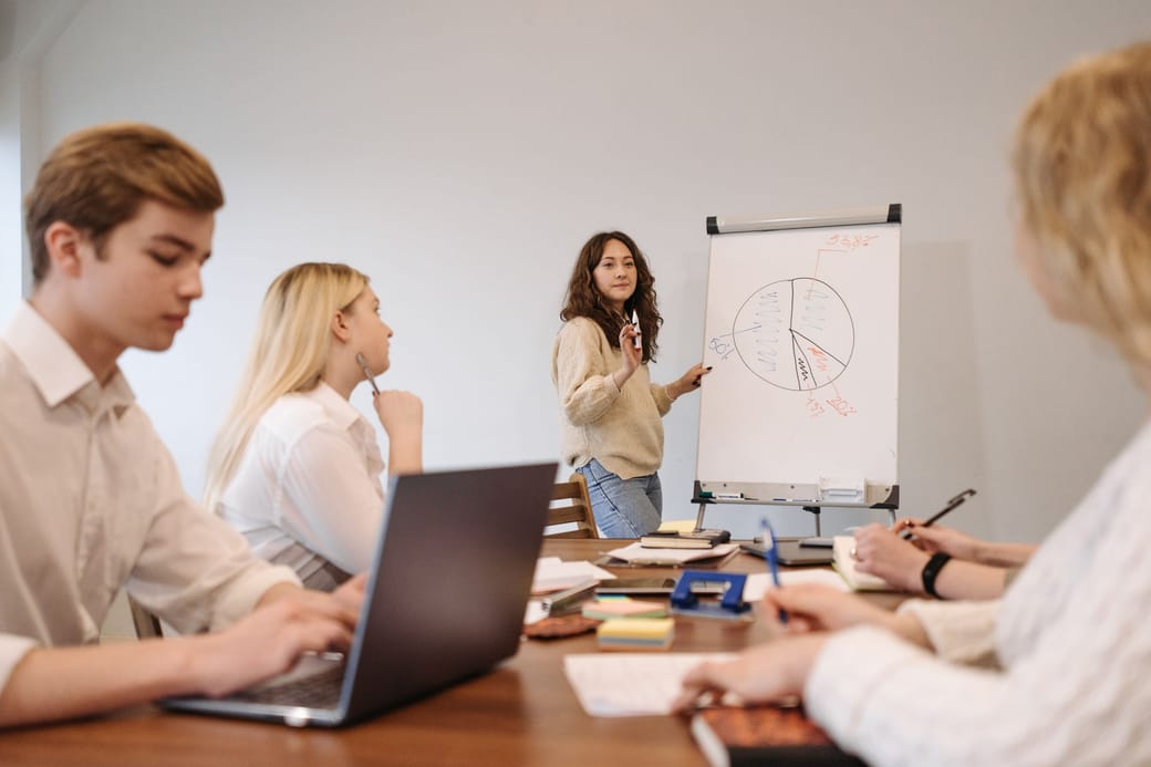 A woman presenting a pie chart on a flip chart to colleagues seated at a table with a laptop and notes