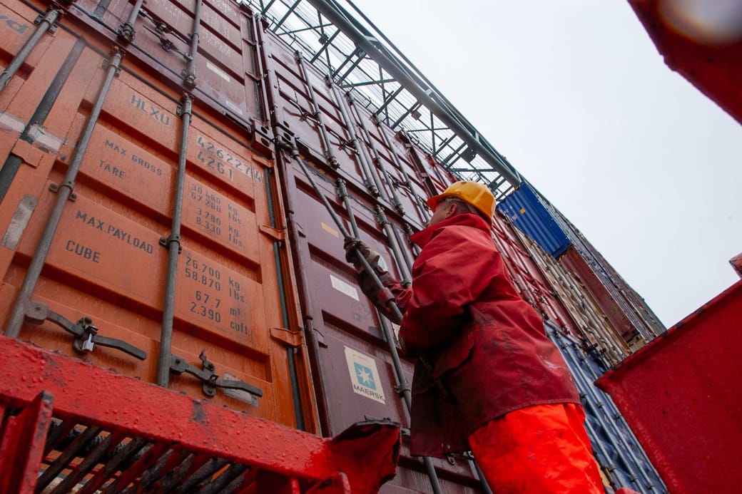 A port worker in a red rain jacket and yellow hard hat looking up at a wall of stacked shipping containers