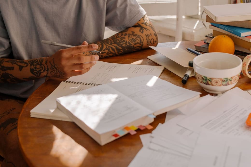 A person at a wooden desk working through an open study guide, with sticky-note tabs, a notebook and loose printed pages