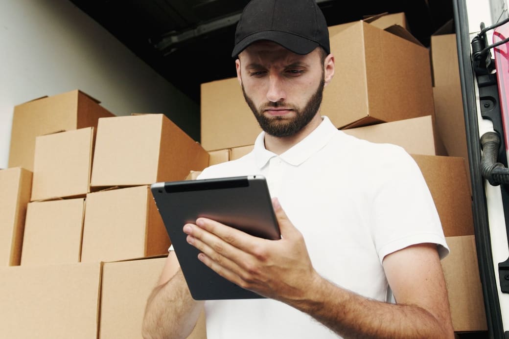 A man in a white polo shirt and black cap checking a delivery list on a tablet in front of a van stacked with cardboard boxes