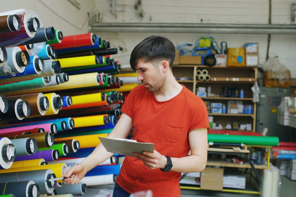 A man in an orange T-shirt holding a clipboard and checking labeled rolls of colored vinyl on a rack in a stockroom