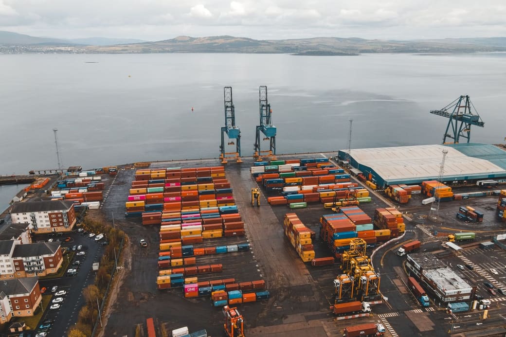 An aerial view of a container port on a wide estuary, with stacked containers, quay cranes and hills beyond