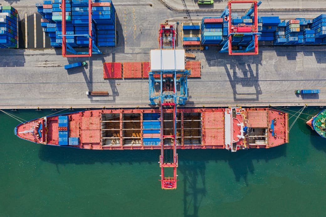 An aerial view of a container ship alongside a port terminal, with gantry cranes and stacked containers on the quay