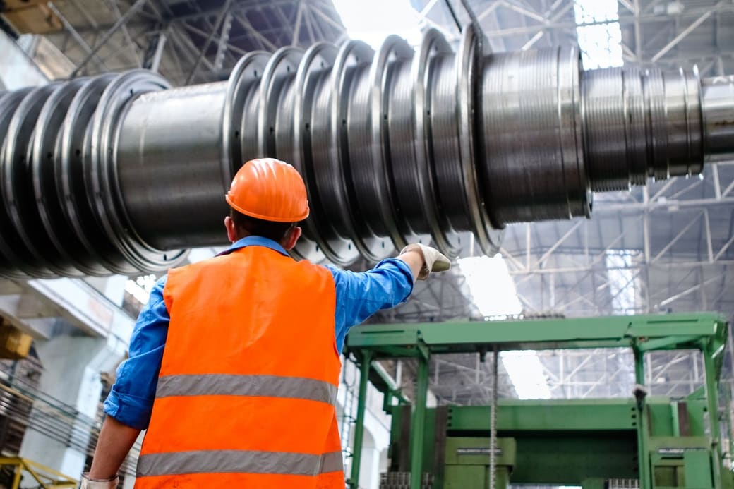 A plant worker in a hard hat and orange vest pointing up at a large machined steel shaft in a factory hall