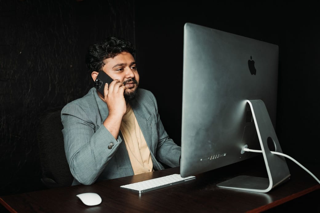 A man in a gray blazer on the phone at a desk, looking at a large computer monitor