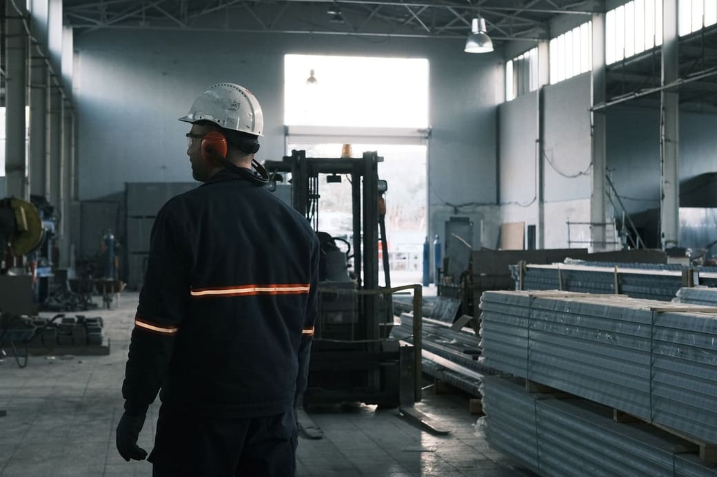 A plant worker in a hard hat and reflective jacket walking the floor of a factory hall past a forklift and stacked steel