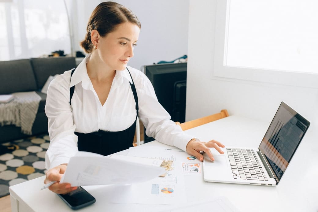A woman at a bright desk comparing a printed report with the charts on her laptop screen