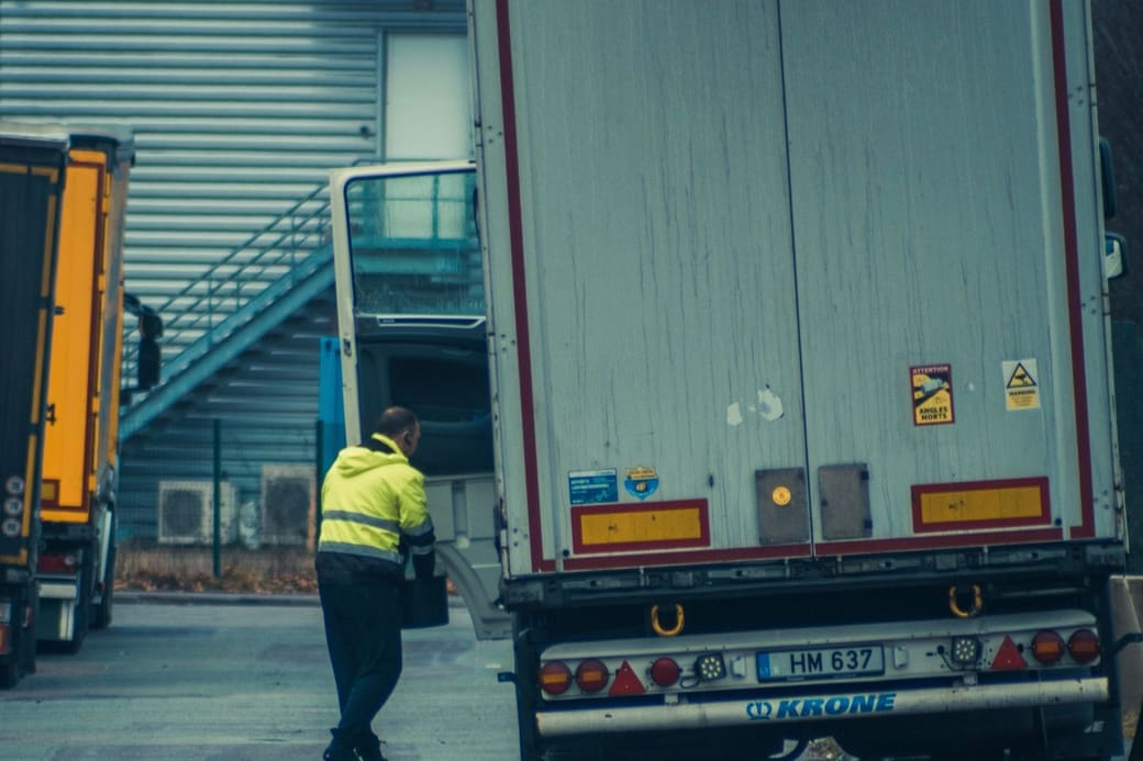 A worker in a high-visibility jacket at the cab of a trailer parked at a loading area outside a distribution building