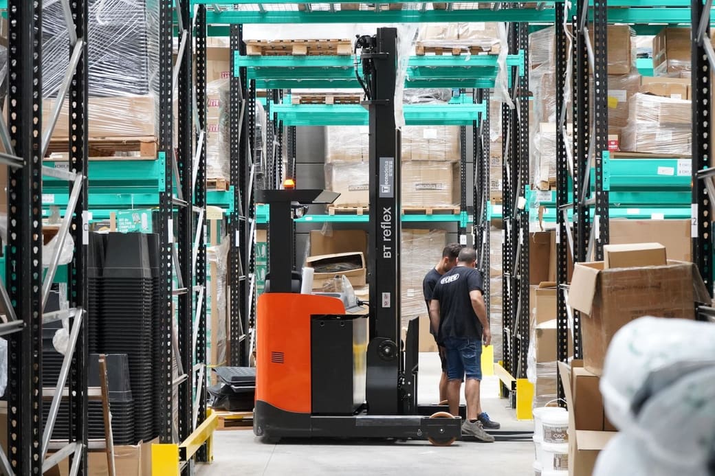 Two warehouse staff beside a reach truck in a racked aisle stacked with boxed stock