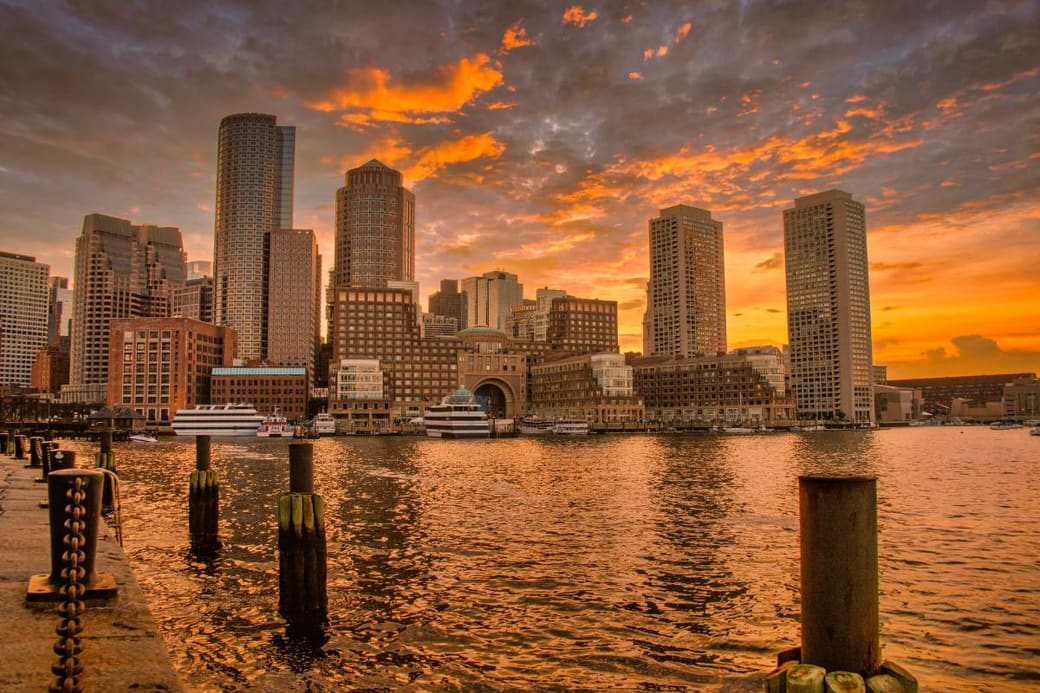 A harbor skyline silhouetted against an orange sunset, reflected in the water