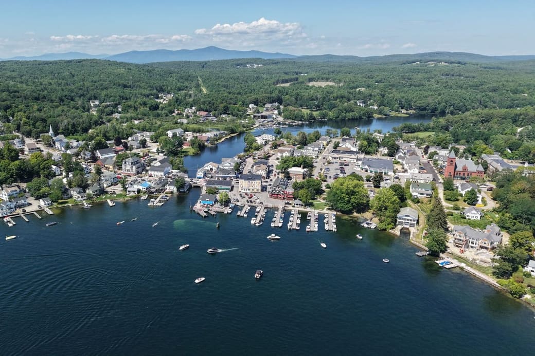 A lakeside town seen from the air, boats moored offshore and wooded hills behind