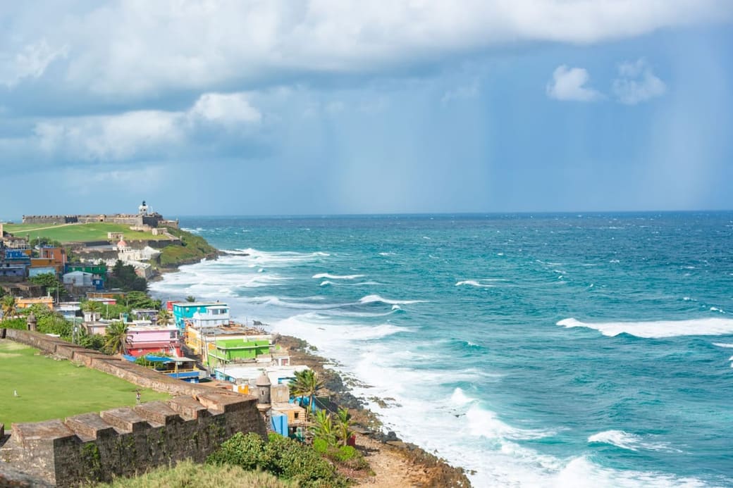 Surf breaking against a rocky shoreline below pastel buildings on a headland